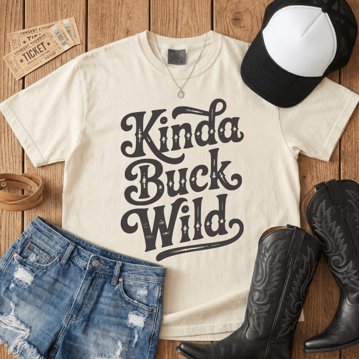 Beige t-shirt with 'Kinda Buck Wild' text, denim shorts, cowboy boots, and a cap on a wooden surface.