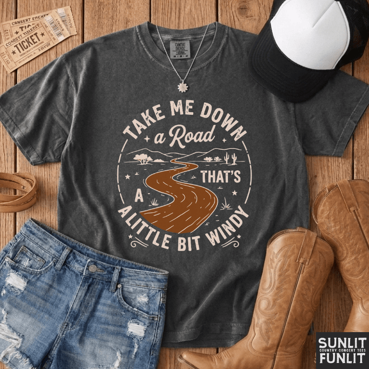 A dark gray graphic t-shirt featuring a western-style design of a winding road through a landscape with the text 'TAKE ME DOWN a Road THAT'S A LITTLE BIT WINDY'. The shirt is styled in a flat-lay arrangement on a wooden background with denim shorts, cowboy boots, a baseball cap, and a ticket.