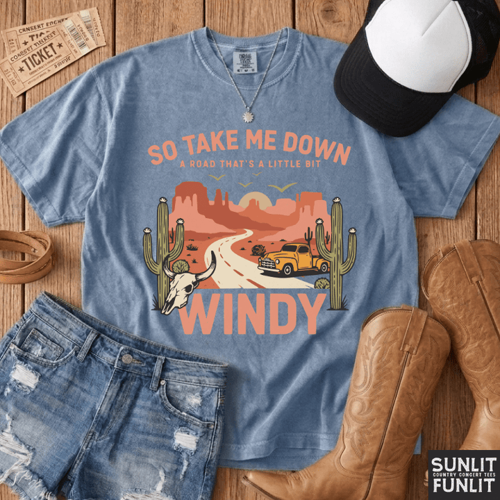 A styled flat-lay of a light blue graphic t-shirt with a desert road trip print, paired with distressed denim shorts, brown cowboy boots, a black and white baseball cap, and a leather belt on a wooden background. The brand logo 'SUNLIT FUNLIT' is visible in the bottom right corner.