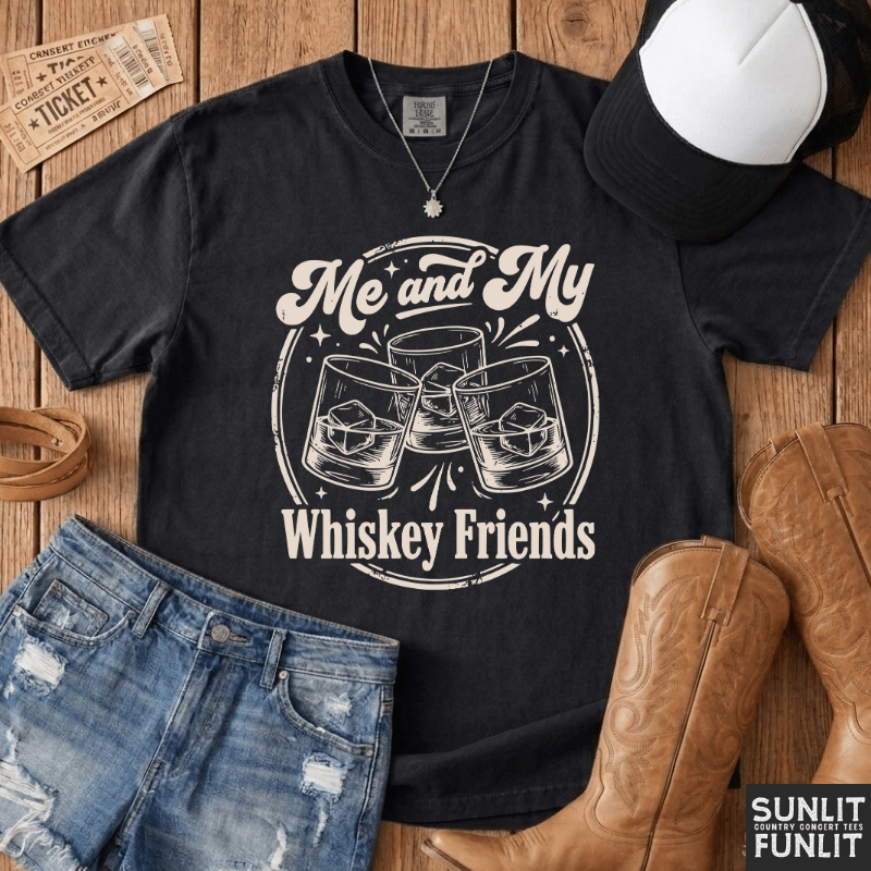A black graphic t-shirt with a white 'Me and My Whiskey Friends' print, styled in a flat-lay with denim shorts, cowboy boots, a baseball cap, and a vintage ticket on a wooden background.