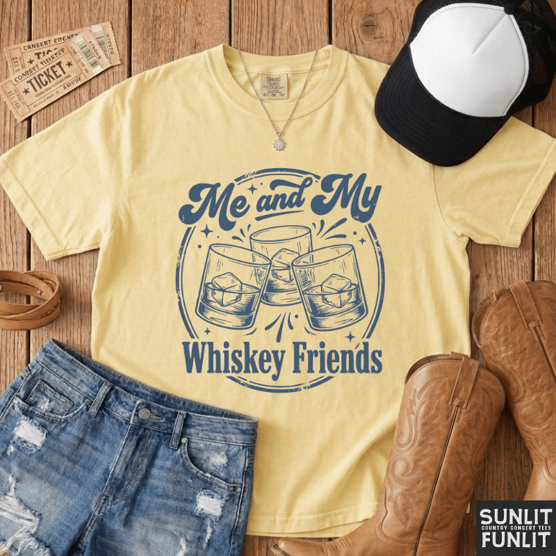 A pale yellow graphic T-shirt laid flat on a rustic wood surface, styled with denim shorts, a black-and-white cap, brown cowboy boots, and a necklace. The shirt reads 'Me and My Whiskey Friends' with an illustration of three clinking whiskey glasses.