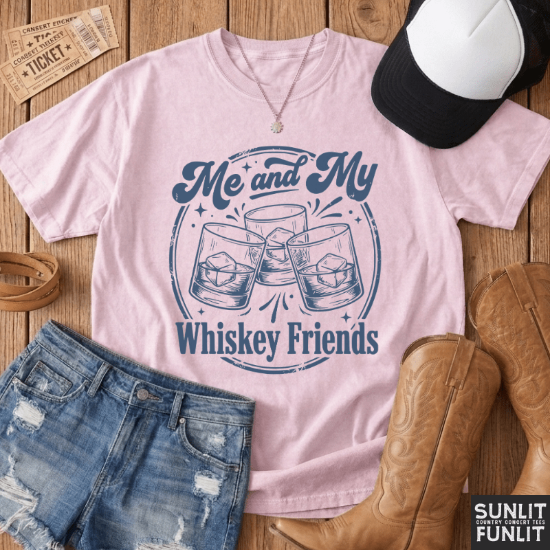 A light pink graphic t-shirt featuring the text 'Me and My Whiskey Friends' in navy blue, styled in a flat-lay arrangement with denim shorts, cowboy boots, a baseball cap, and a necklace on a wooden background.