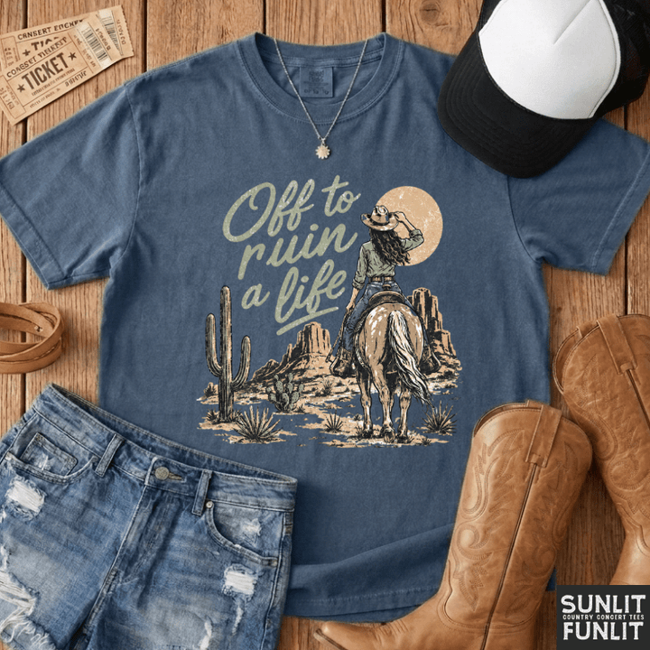 A styled flat-lay of a blue graphic T-shirt on a wood surface, surrounded by jeans, cowboy boots, a cap, and tickets. The shirt reads 'Off to ruin a life' with a western rider, cactus, and moon graphic.
