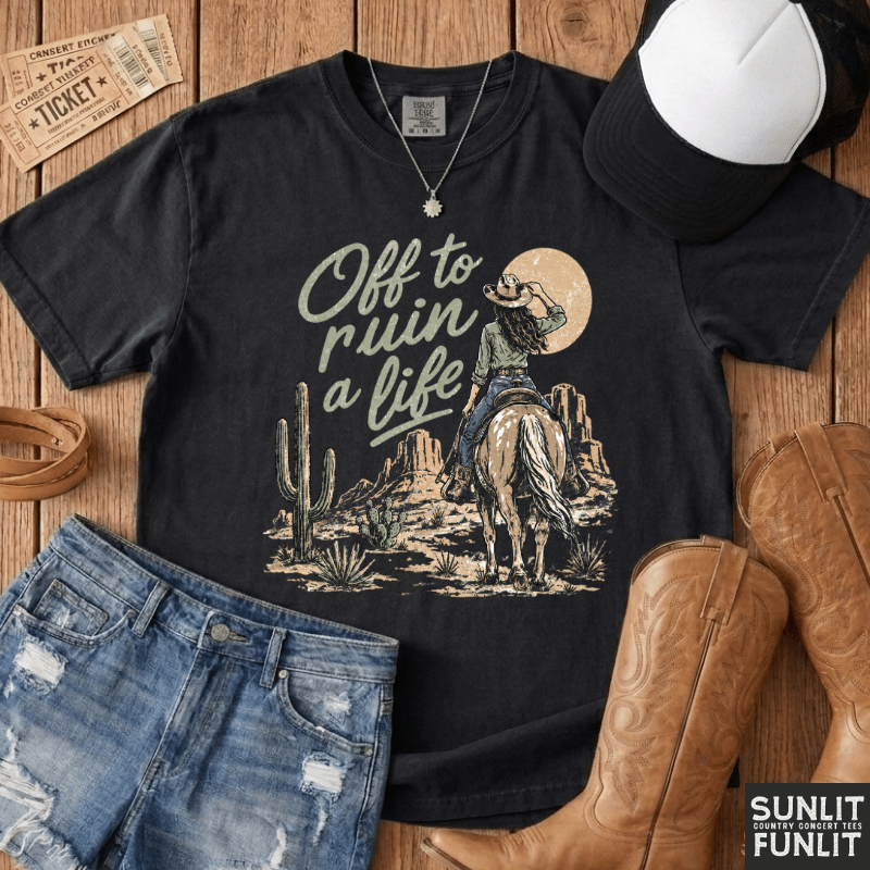 A black graphic t-shirt featuring a western cowboy design with the text 'Off to ruin a life' and a desert landscape, styled in a flat lay with denim shorts, cowboy boots, a baseball cap, and a necklace on a wooden background.