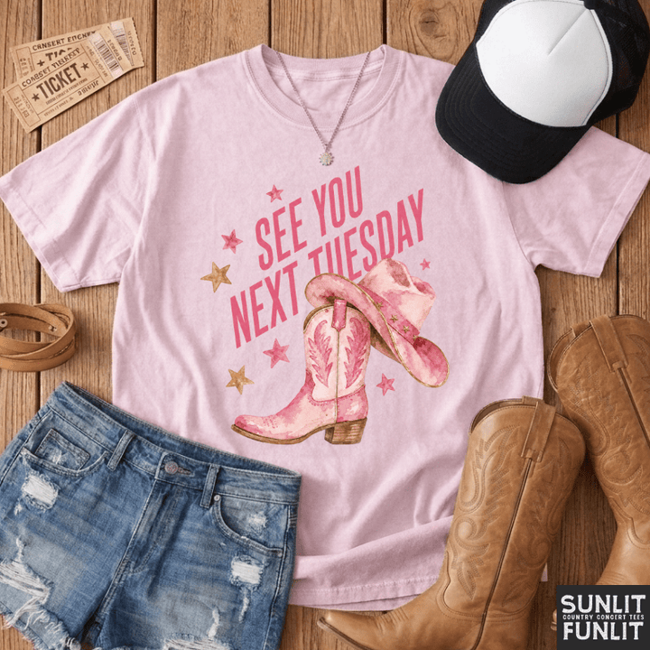 A light pink graphic t-shirt featuring the text 'SEE YOU NEXT TUESDAY' in pink with cowboy boots and stars, styled in a flat lay with denim shorts, a black and white cap, brown cowboy boots, and tickets on a wooden background. The brand logo 'SUNLIT WITH FUNLIT' is visible in the bottom right corner.