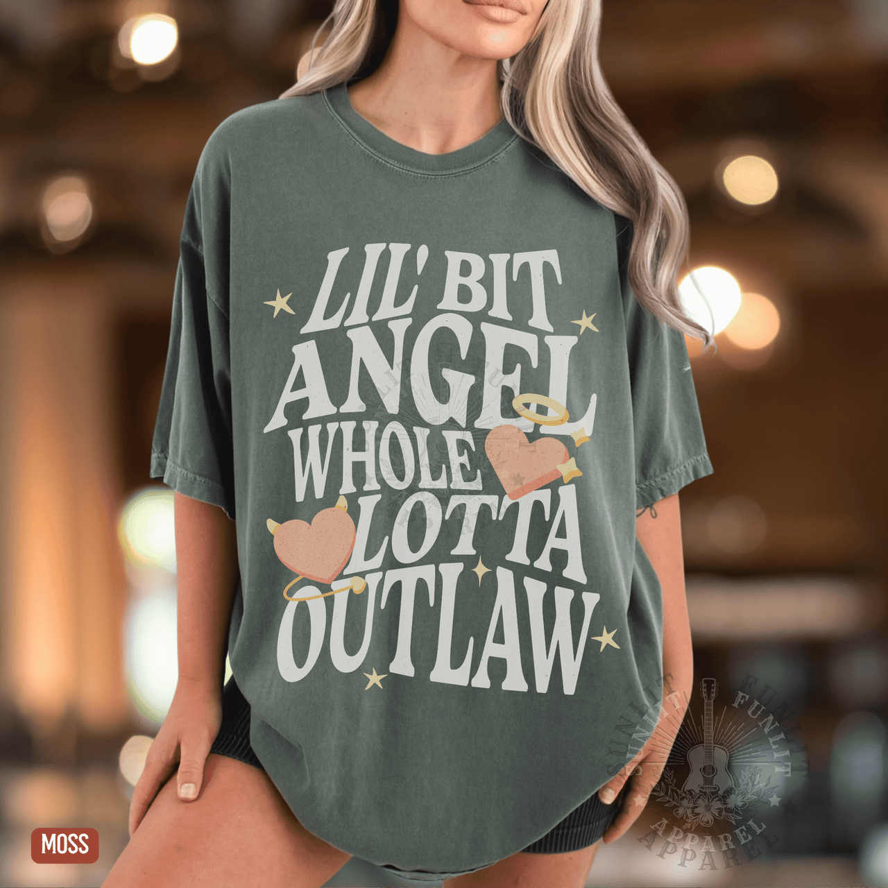 A woman wearing a green t-shirt with the text "Lil' Bit Angel Whole Lotta Outlaw" printed on it.