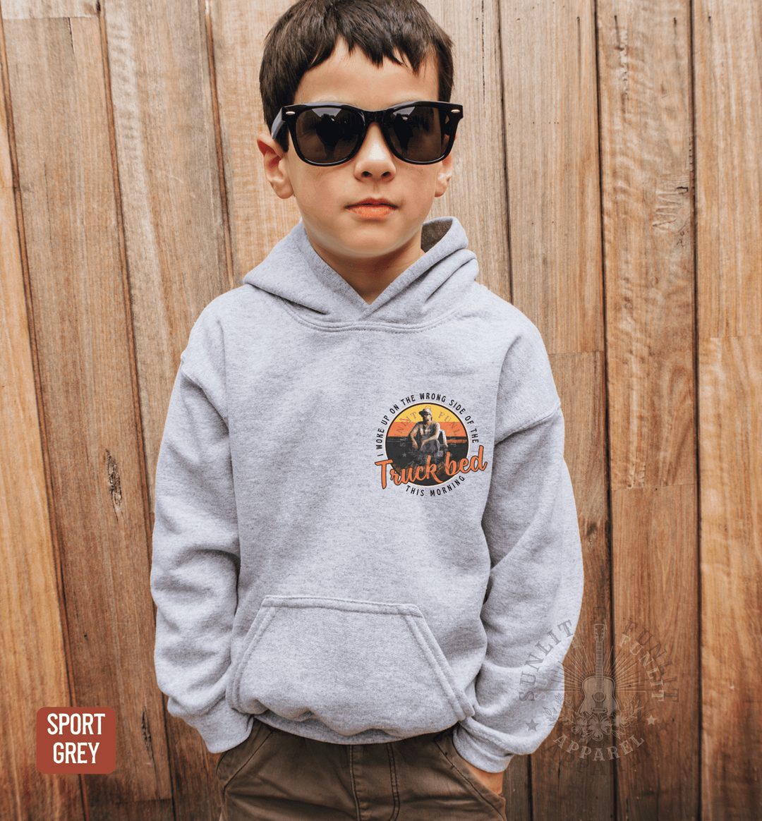 A young boy wearing a grey hoodie with a graphic design and sunglasses stands in front of a wooden fence.